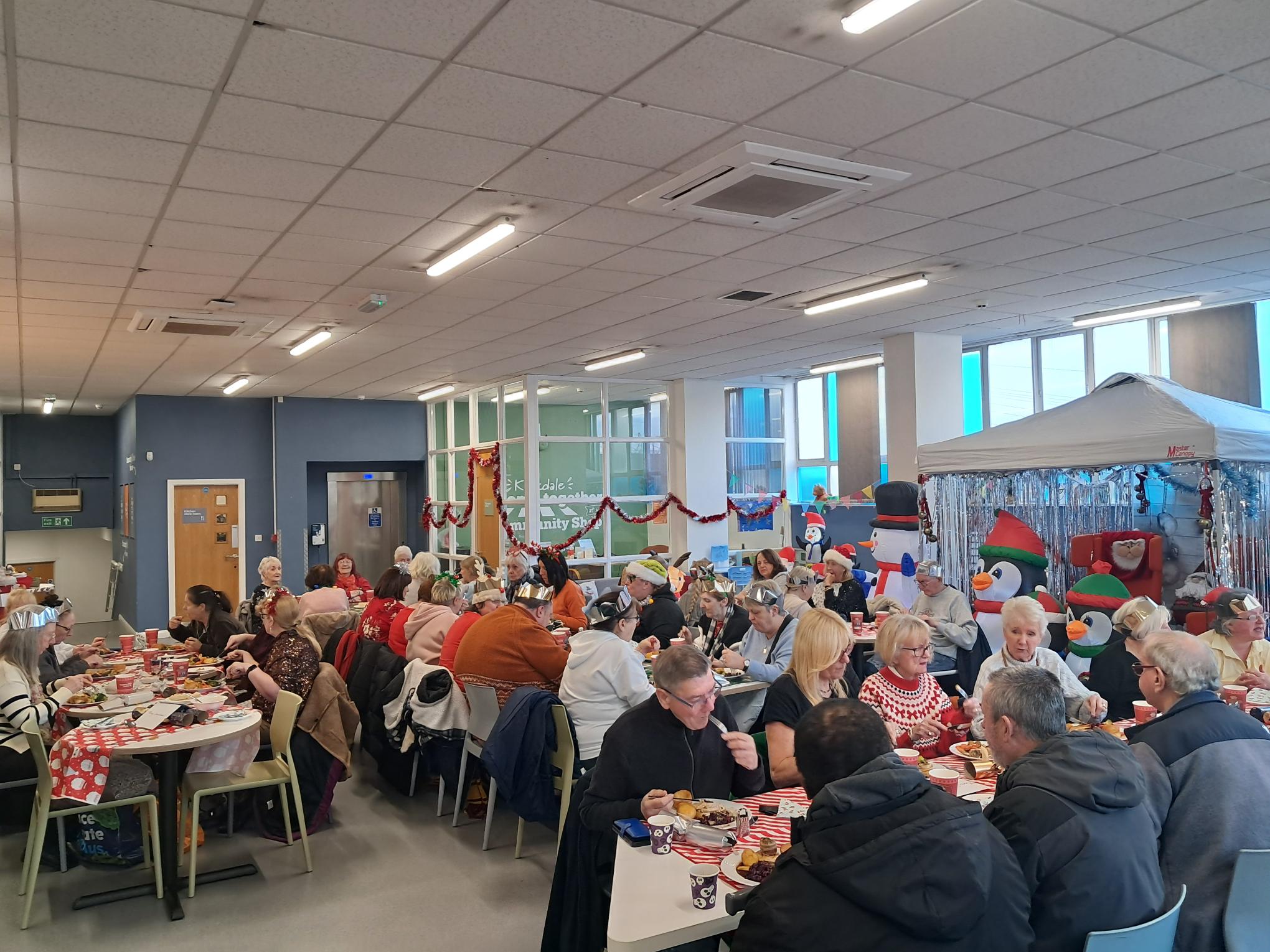 Multiple people, wearing Christmas hats and jumpers sat at tables eating Christmas dinner in a room with Christmas decorations